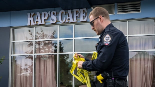 A Surrey Police officer removes tape outside of Kap's Cafe, owned by Bollywood celebrity Kapil Sharma, in Surrey, B.C., on July 10, 2025. The Surrey Police Service (SPS) says it was called to Kap’s Café on 120 Street near 84 Avenue around 1:30 a.m. Thursday, after shots were fired into the business. A police officer rolls up yellow tape outside a business that says Kap's Cafe in white lettering against a blue background.