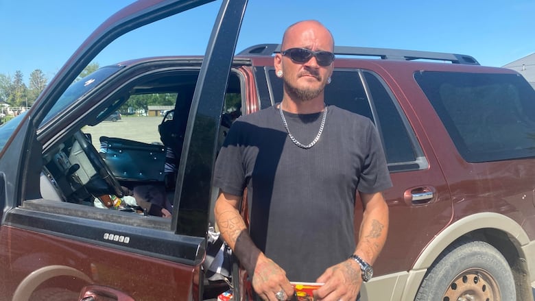 A man wearing a chain, black T-shirt and sunglasses stands in front of a dark car.