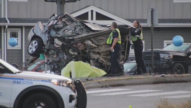 A damaged car is pictured with its front crashed into a highway divider and back wheels in the air. Two police officers stand in front.