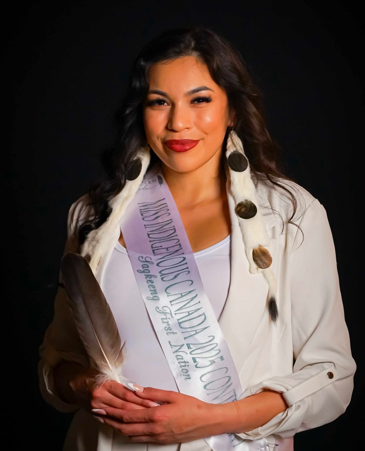 A woman with long black hair, wearing a white blazer and t shirt and a sash that reads 'Miss Indigenous Canada 2025 Contestant' smiles into the camera.