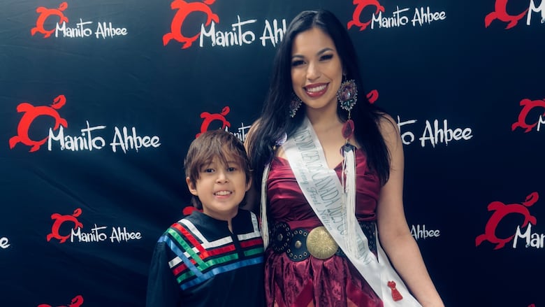 A woman with long dark hair, wearing a maroon dress with a sash that reads "Miss Indigenous Canada 2025 Contestant", stans and smiles next to a young boy with short black hair, wearing a dark blue shirt with multi-coloured ribbons on it.