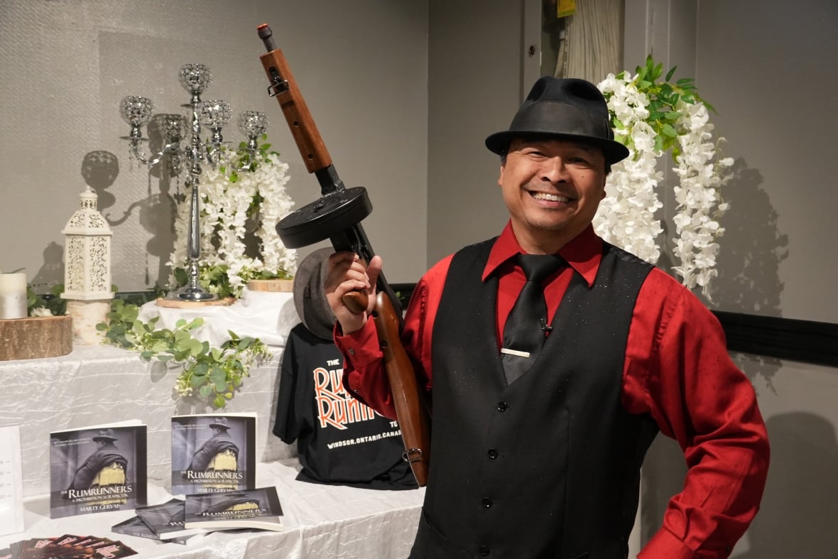 A man is wearing a black fedora hat and a 1920s era red and black vest suit. He is posing with a prop tommy gun in front of a merchandise table.