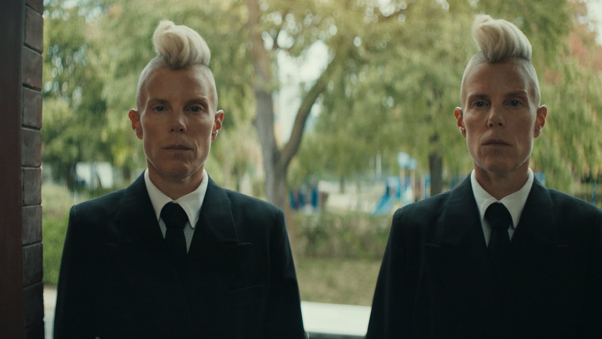 Twins, wearing a black suit, with blonde mohawks stand at a doorway.