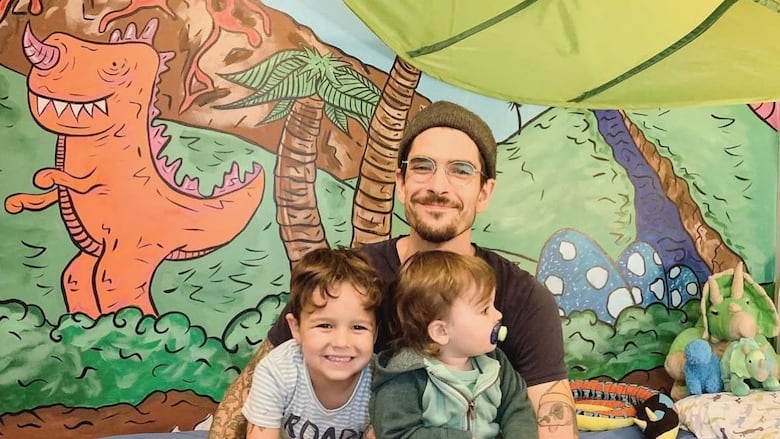 A man and two children sit in front of a dinosaur mural.