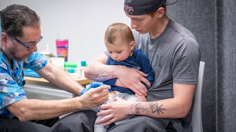 A man holds a baby on his lap. Another man, a healthcare provider wearing blue camouflage scrubs sits across from them. He is vaccinating the child on his thigh. The baby looks down, intently at the shot.