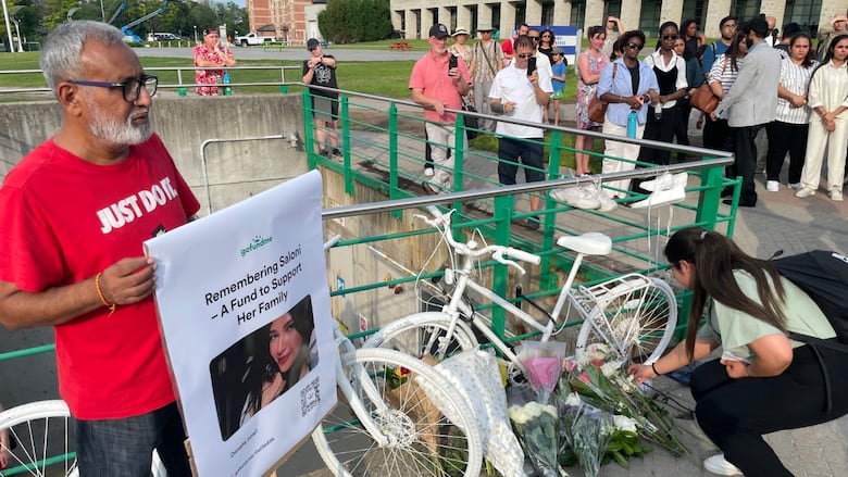 A man holds a fundraising sign to support the family of a woman who died, next to flowers and white bicycles representing dead cyclists.