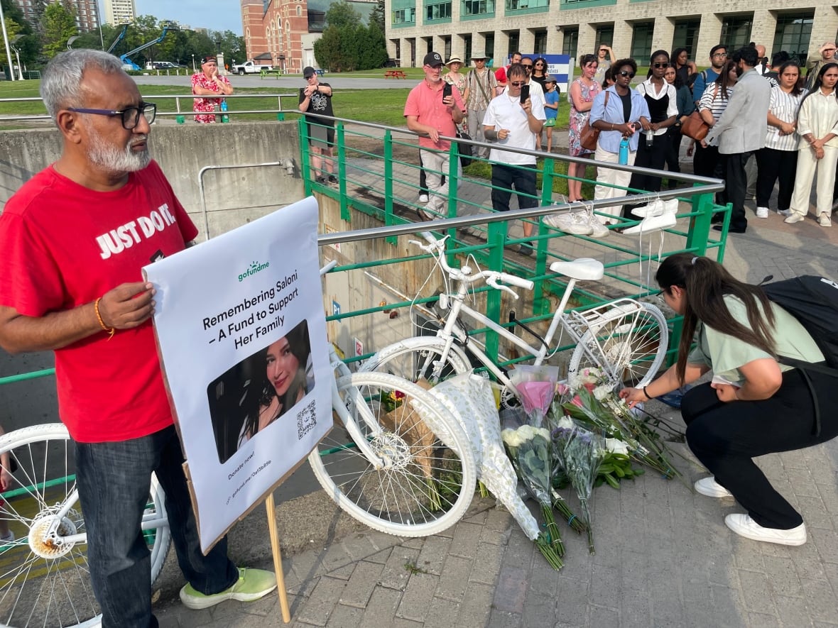 A man holds a fundraising sign to support the family of a woman who died, next to flowers and white bicycles representing dead cyclists.