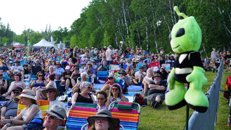 Crowd of people sitting on grass, with stuffed green alien sitting on a fencepost in the foreground
