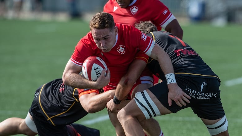 A rugby player tries to break through two players with the ball in his right hand.