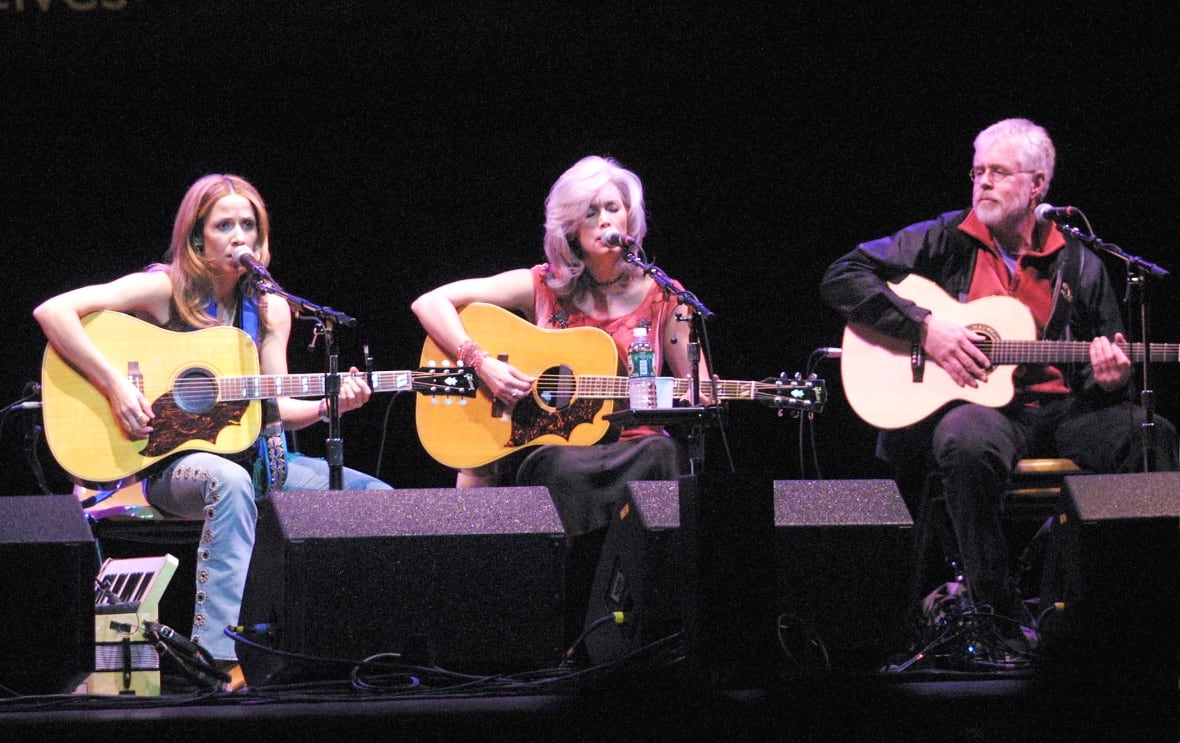 Three musicians with guitars are shown on stage. Two are women and one is a man.
