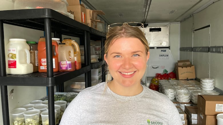 A woman in a gray t-shirt stands in the back of a cooled truck, behind her food and food products in packages and boxes can be seen.