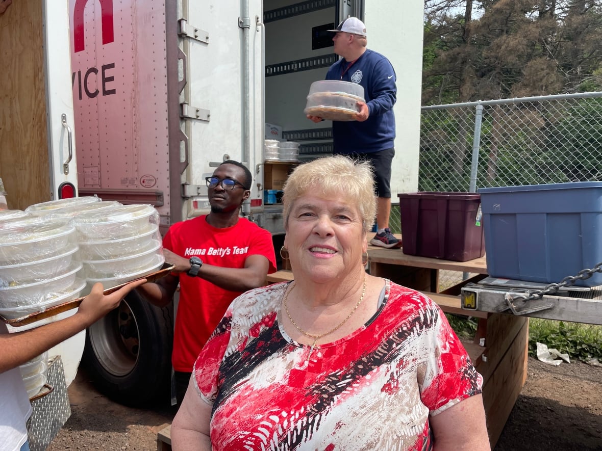 A woman in a red, white and black designed shirt stands behind two trucks, three people can be seen transferring packaged food into the charities truck.