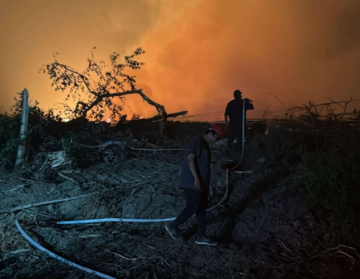 Dark orange sky willed with smoke with shadows of two firefighters carrying a hose in the foreground