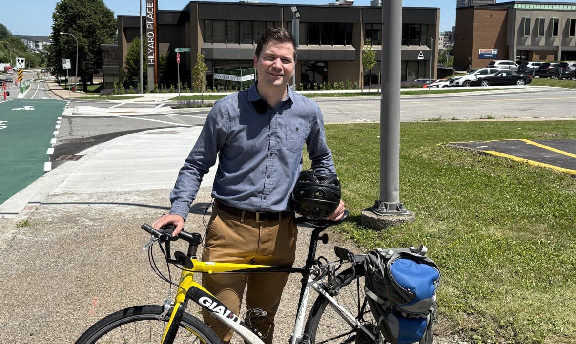 Man standing with a bicycle held in front of him and a helmet under his arm smiling at camera on a sunny day.