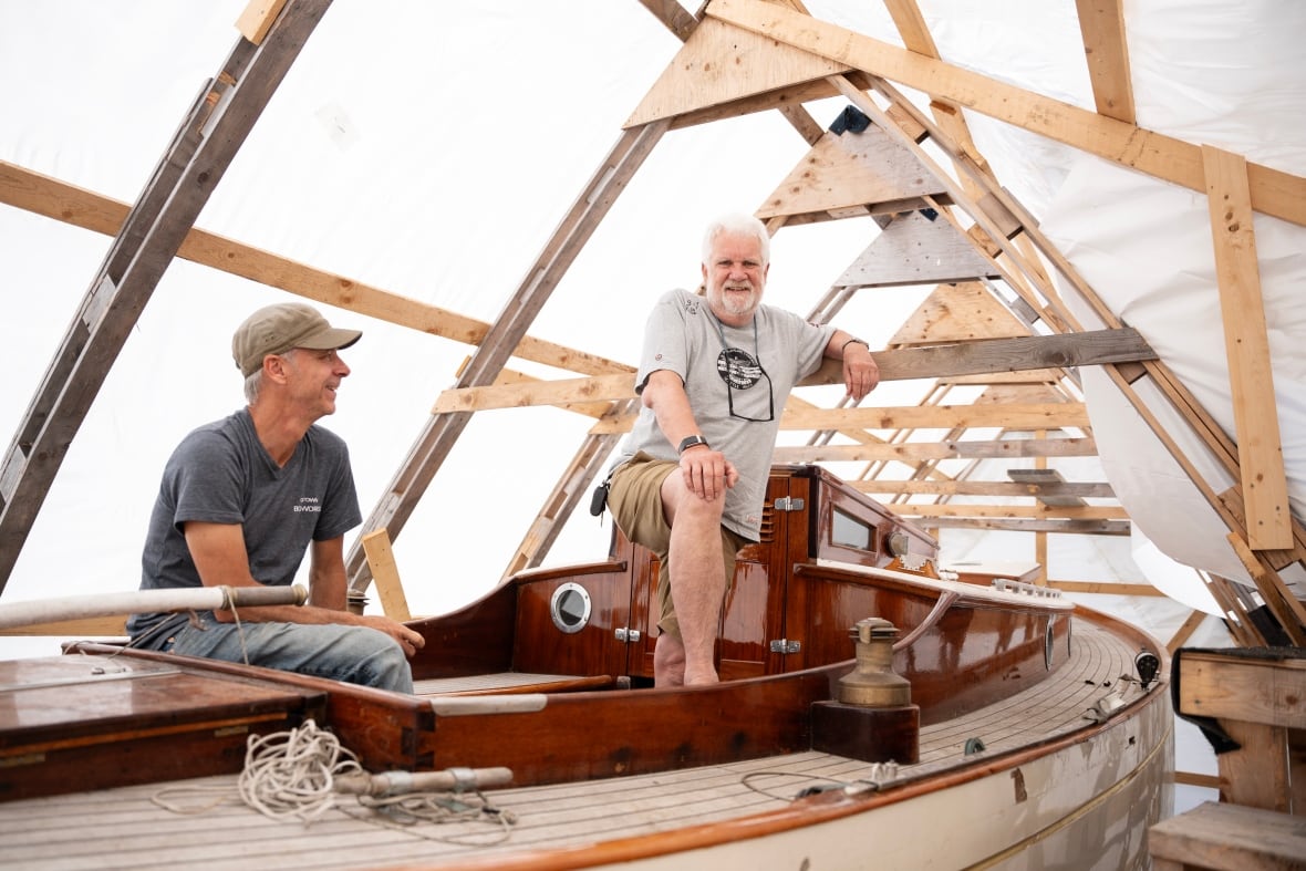 One person sits in and one person stands in a boat that's inside a tent.