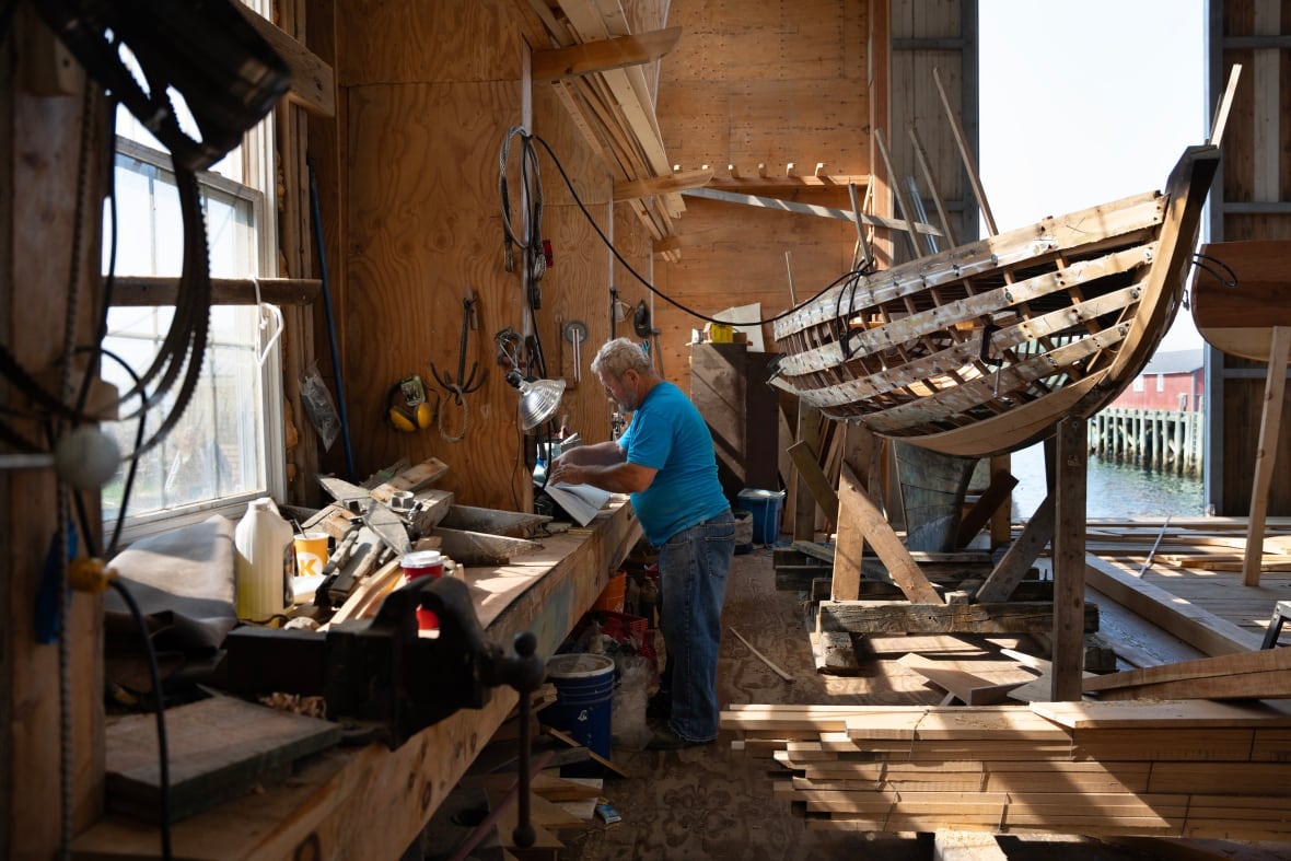 A man stands at a workbench. A boat is under construction in the background.