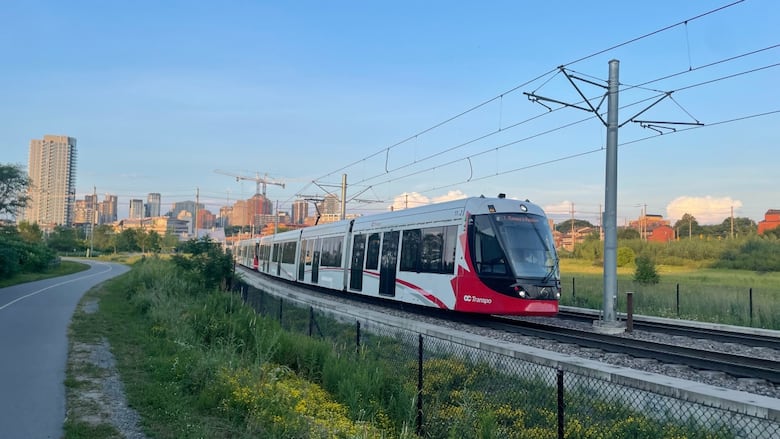 A red and white light rail train moves between stations on a summer evening.