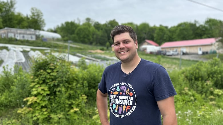 A young man with short brown hair brushed back and to the side smiles toward the camera for an upper body portrait with one hand on his hip. In the background is a hillside with garden fields and a few single story wooden structures.