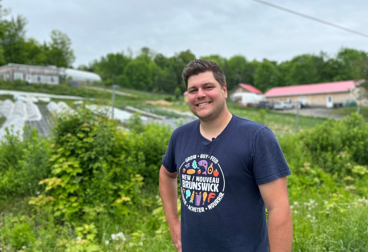 A young man with short brown hair brushed back and to the side smiles toward the camera for an upper body portrait with one hand on his hip. In the background is a hillside with garden fields and a few single story wooden structures.