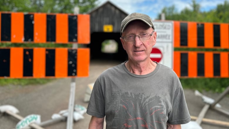 A white man wearing blown hat and glasses standing in front of a closed bridge.