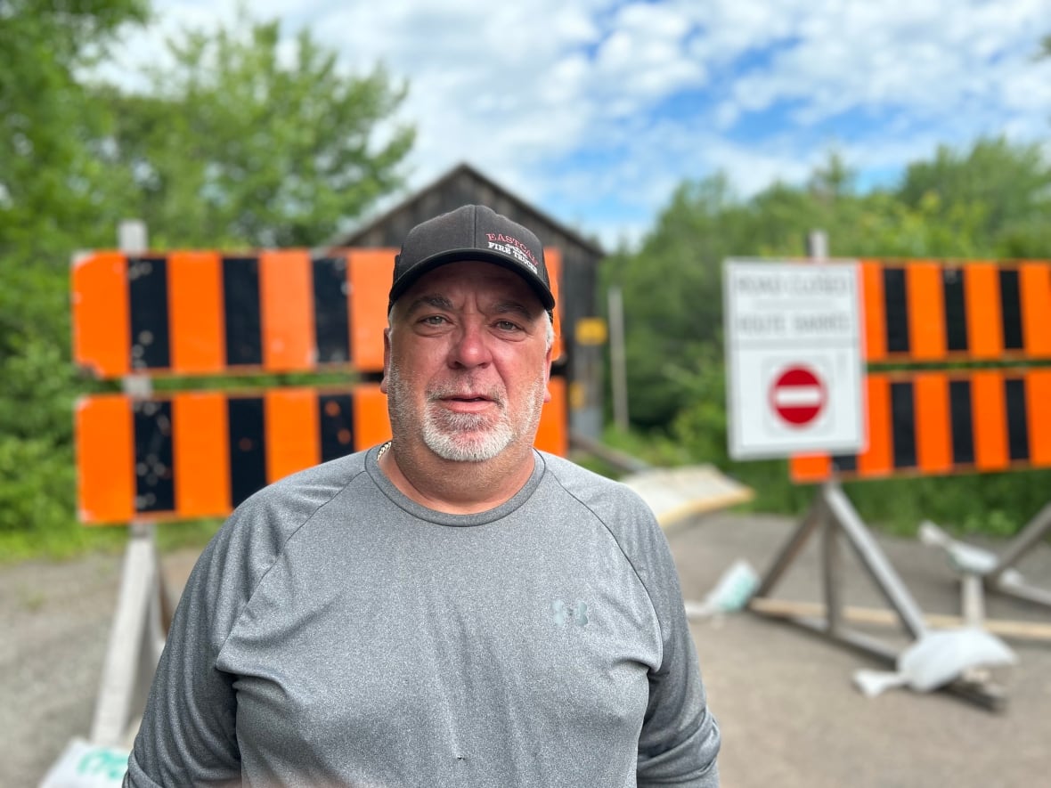 A white man with white beard. He is wearing a hat and standing in front of a closed bridge.