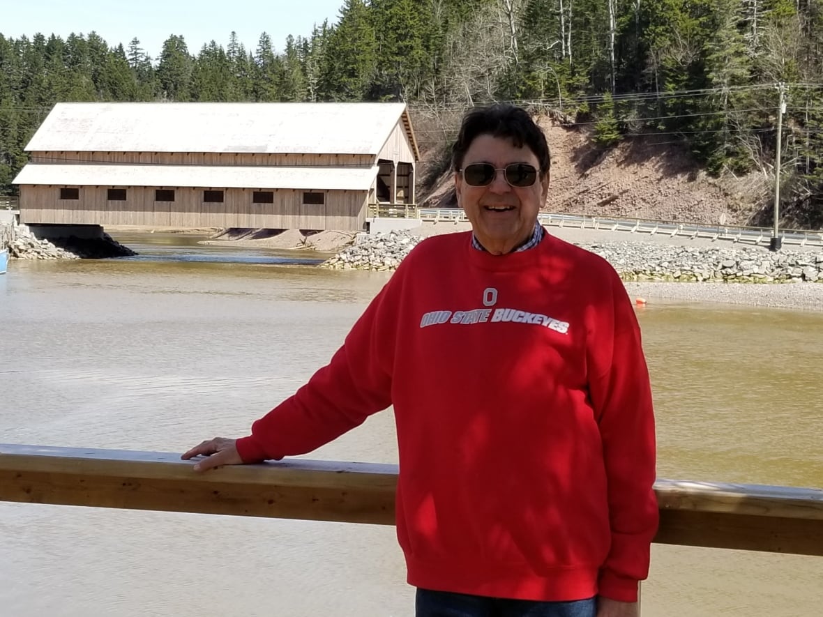 A white man with black hair, wearing shades, standing in front of a wooden covered bridge,
