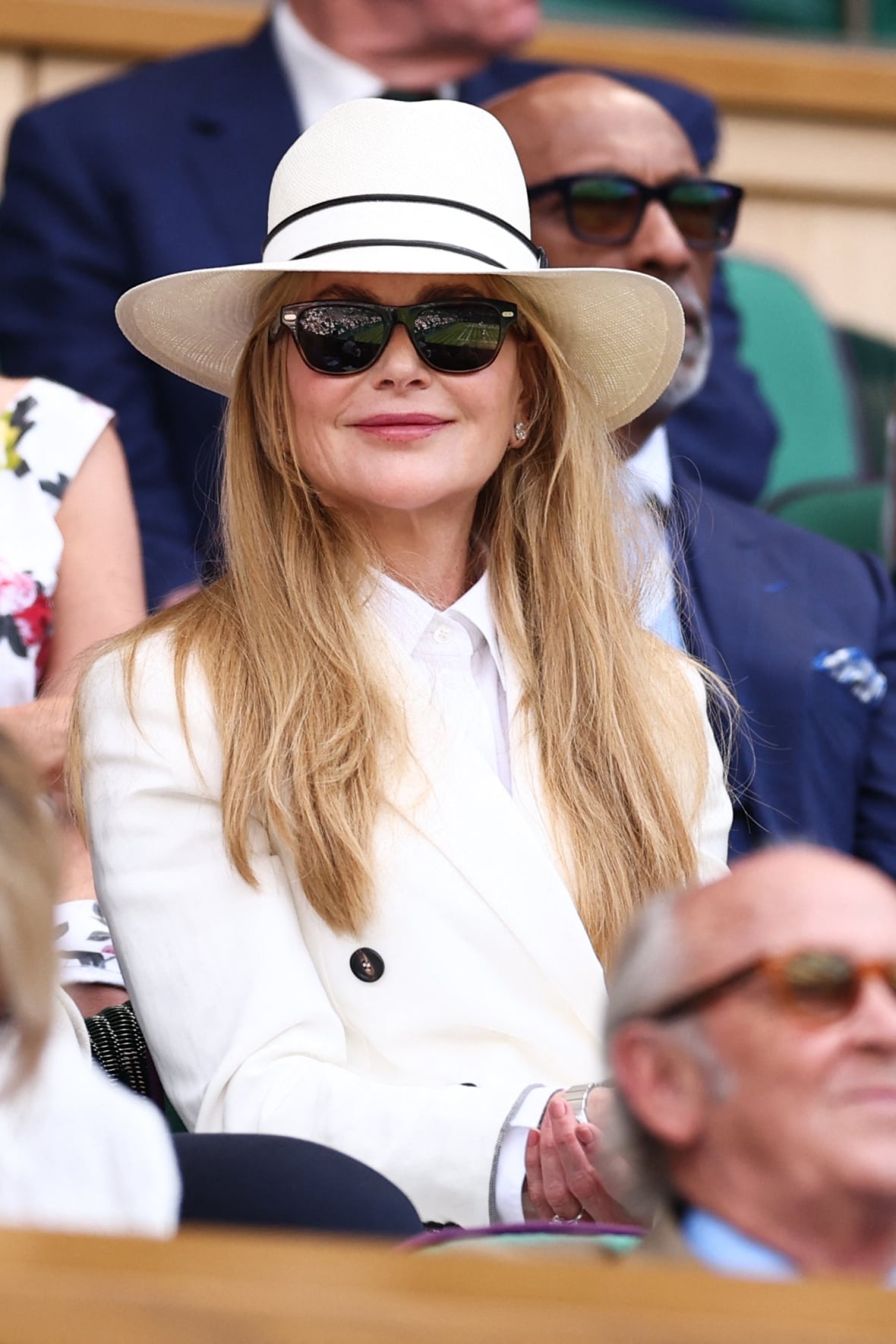 A woman sits in a crowd wearing a white suit, white hat and sunglasses.