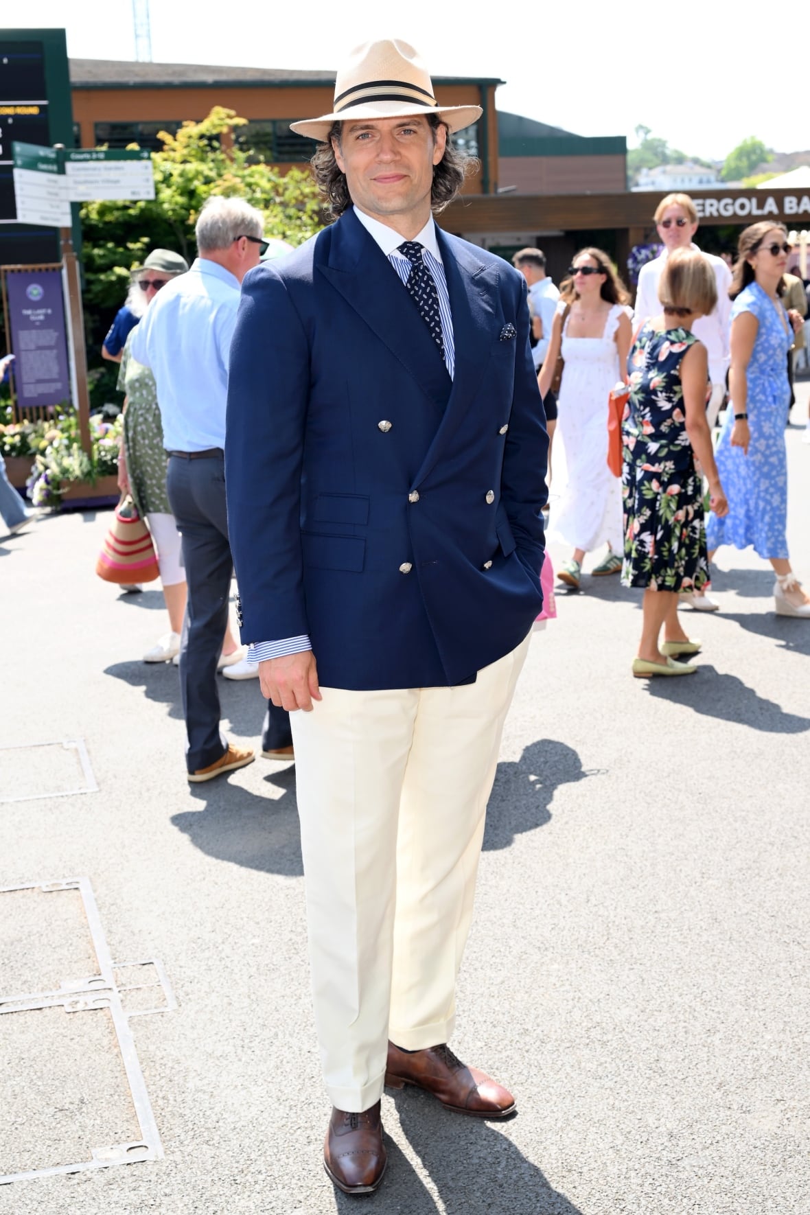 A man poses for a photo wearing a double-breasted navy blazer, white trousers, a striped shirt and a straw hat.
