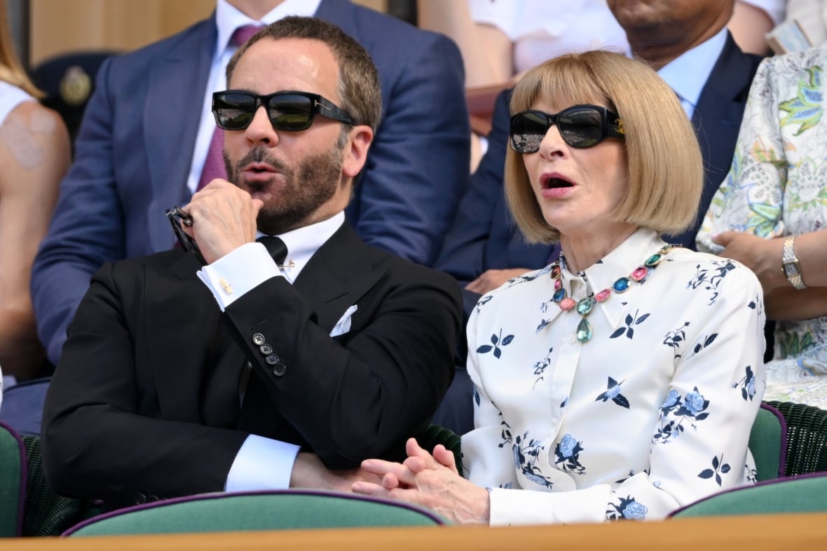 A man and a woman sit in a crowd wearing sunglasses. He's wearing a black suit and tie. She's wearing a white shirtdress with floral pattern.