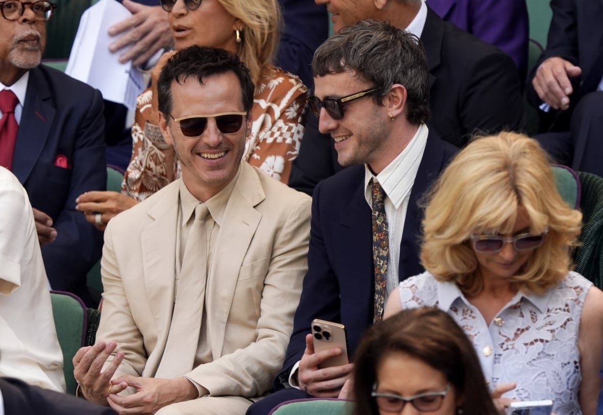 2 men smiling as they watch tennis in a crowd. One is wearing a tan suit, the other is wearing a navy suit.