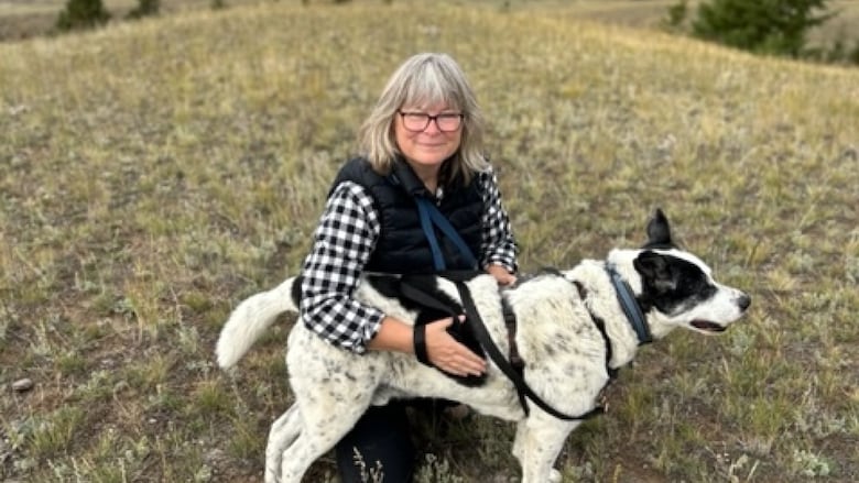 A woman is pictured sitting on a dry, grassy hill with a white and black dog.