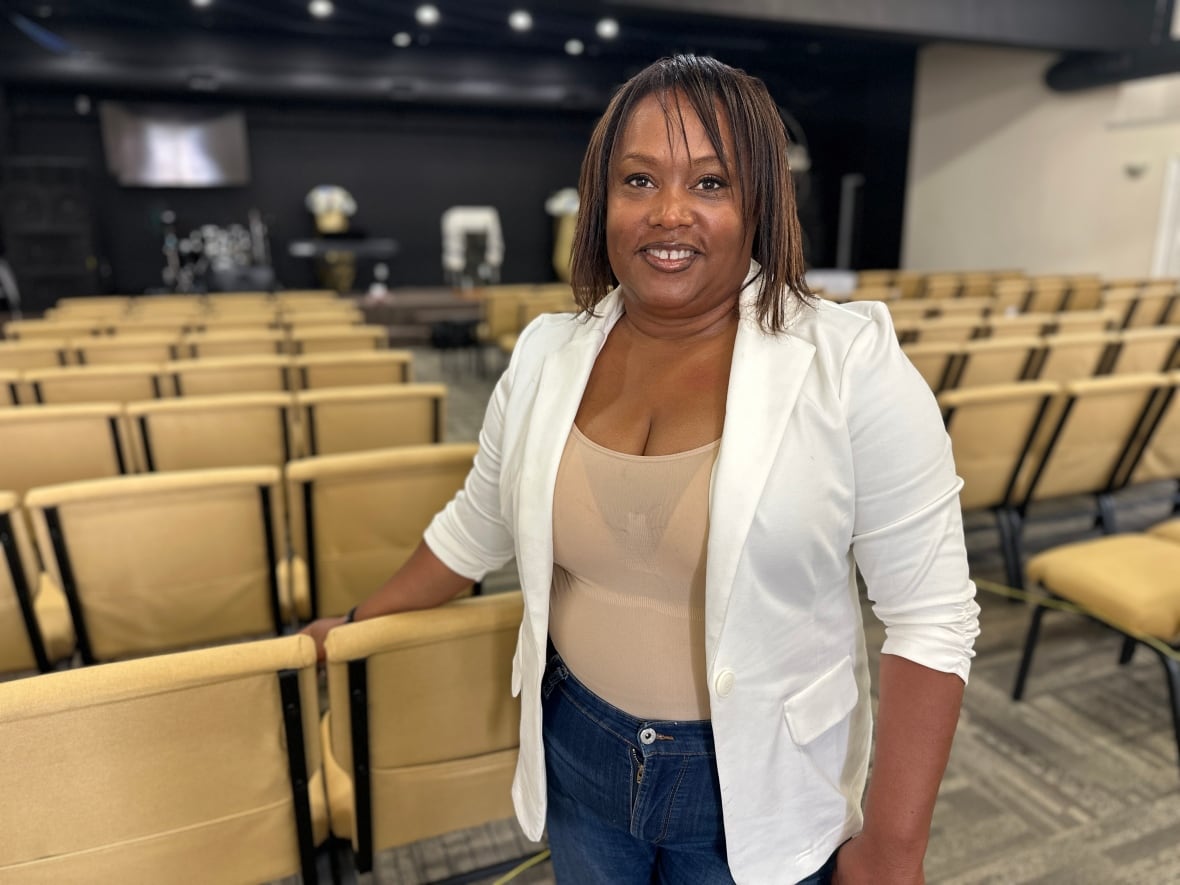 A woman in a white blazer is standing in a large room with rows of brown chairs and a stage in the background. 