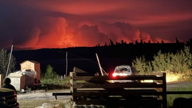 Vehicles are seen shining headlights into houses by a body of water. In the background is a pink and orange sky and large plumes of smoke.