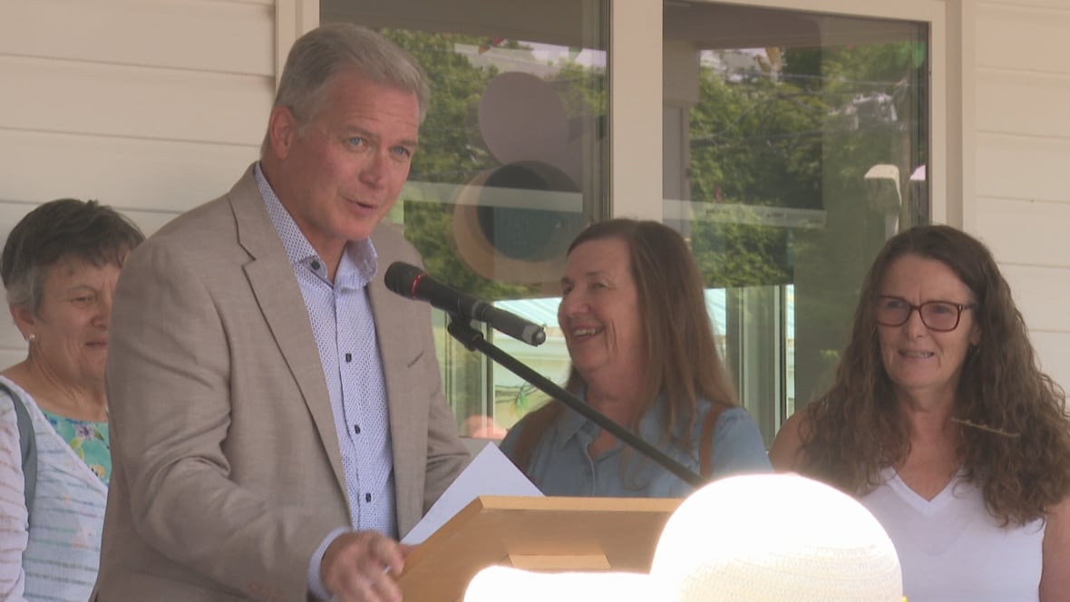 Man in a brown blazer and blue shirt at a podium, with three happy women behind him.