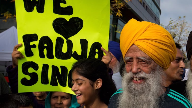 An elderly bearded man with a turban is shown smiling as he stands beside a young girl.