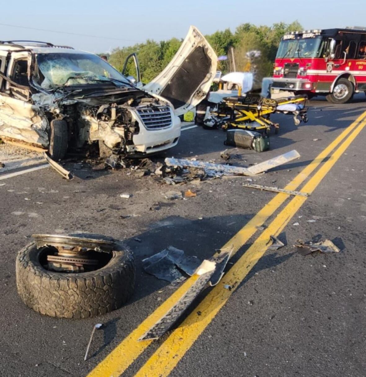 A collision scene with a white van's front end damaged and the passenger side tire is on the roadway. There is debris on the road and a fire truck in the background.