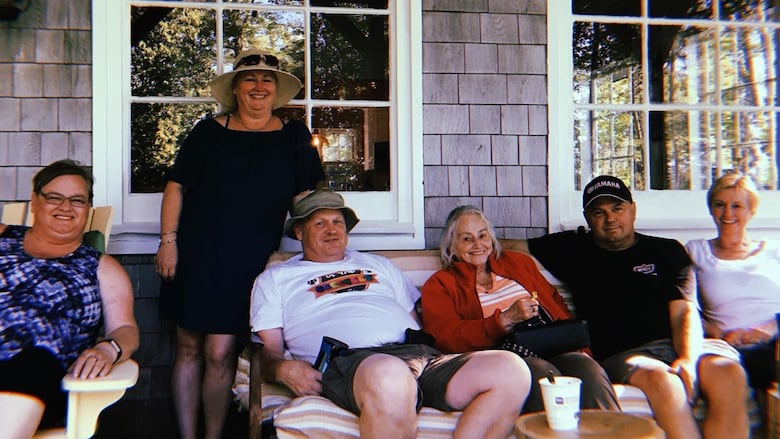 Five people sitting on a porch bench with grey house siding and two windows behind them.