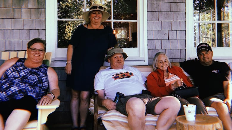 Five people sitting on a porch bench with grey house siding and two windows behind them.