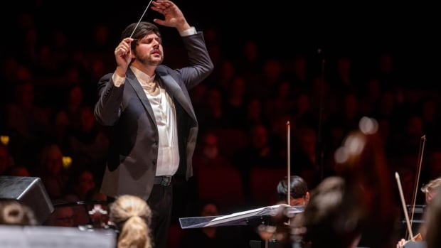 Male conductor leading an orchestra onstage with a focused expression, holding a baton above his head.