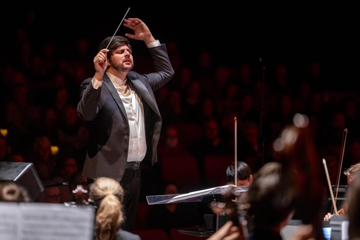 Männlicher Dirigent führt mit konzentriertem Gesichtsausdruck ein Orchester auf die Bühne und hält einen Taktstock über seinem Kopf.