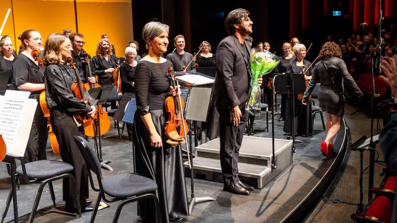 Musicians on stage with a conductor holding a bouquet, smiling at an audience.