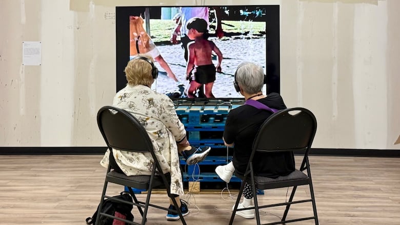 Seen from behind, two women sit on folding chairs, watching a screen sat upon a stack of loading skids.
