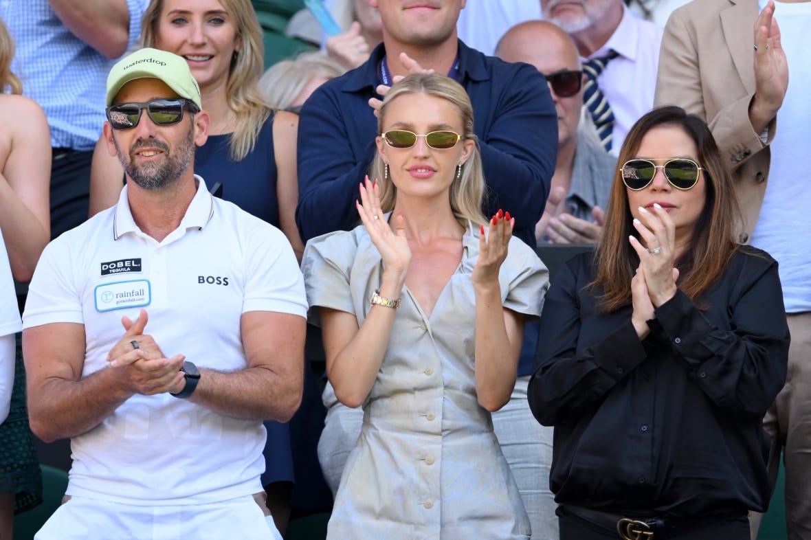 A woman wearing sunglasses and a dress claps as she watches tennis in the audience.