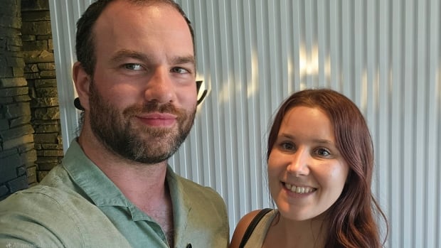 A man and a woman take a selfie. The man has short hair and a beard and is wearing a green collared shirt. The woman has wavy auburn hair and is wearing a tank top. 