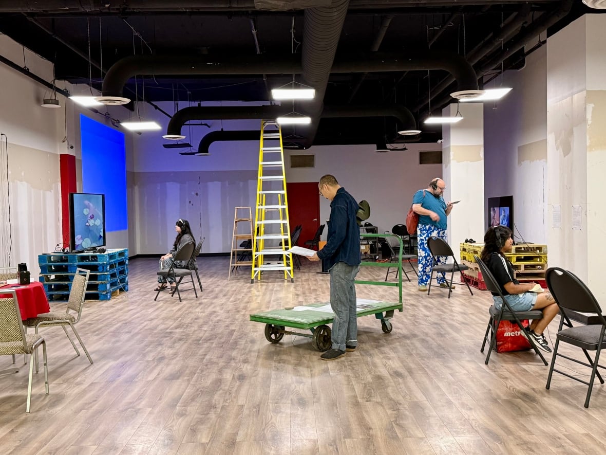 In a mall store emptied of fixtures, people sit on folding chairs, watching videos from screens propped on top of stacks of skids. A dolly sits in the middle of the room.