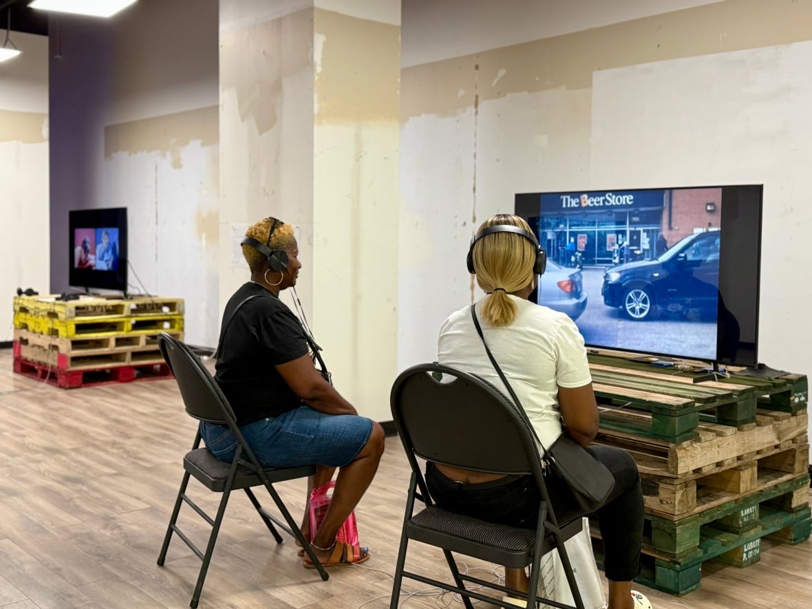 Seen from behind, two people sit on folding chairs, watching a screen placed on top of a stack of loading skids.
