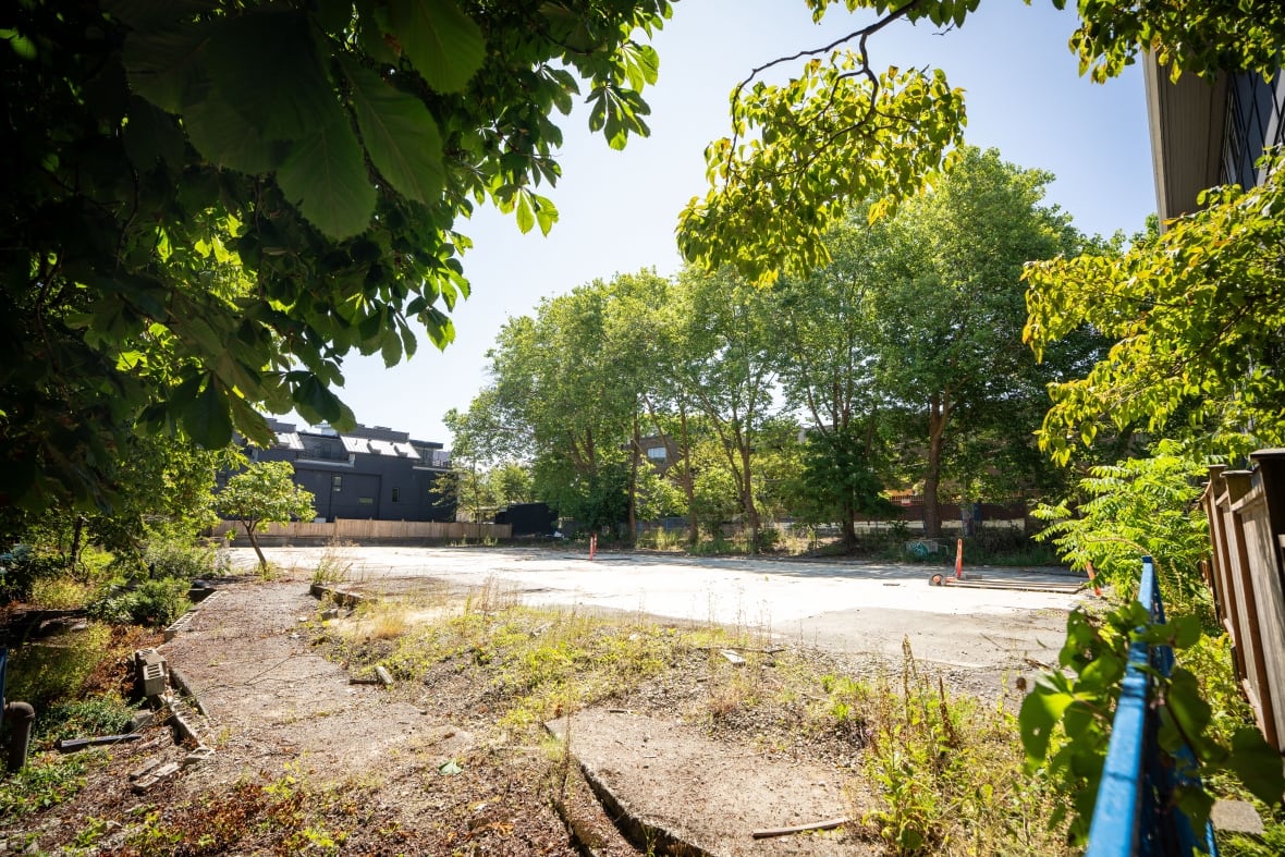 An empty property lot, with some grass and trees surrounding a paved area.