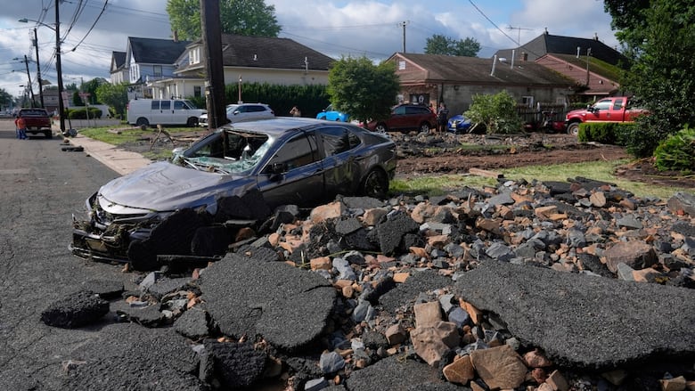 A gray car with its windows smashed sits on top of a pile of debrise under a blue and cloudy sky.
