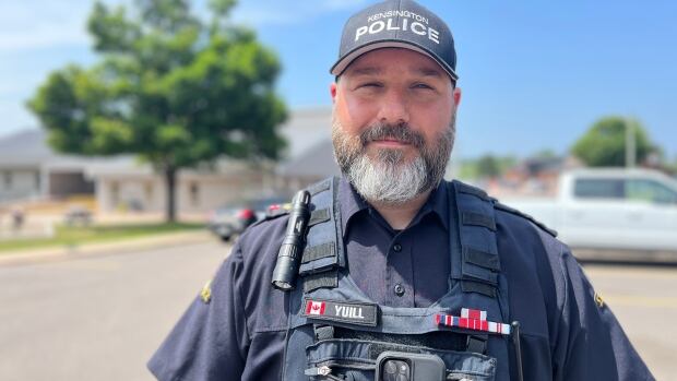 A man with a grey and black beard in police tactical gear standing in front of a building. 