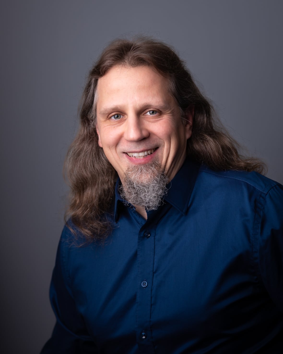 Profile photo of a person looking at the camera, person has long hair and is smiling. He is wearing a blue shirt.